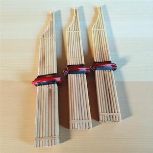 Puede incluir: Tres flautas de pan tradicionales de bambú con una banda roja y negra en el centro. Las flautas de pan están hechas de finas cañas de bambú de color claro y están dispuestas sobre una superficie de madera clara.