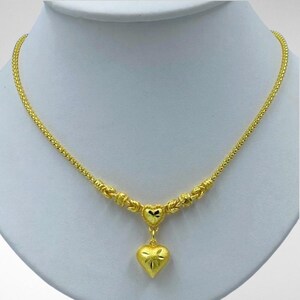 May include: A gold-colored necklace with a heart pendant. The necklace features a delicate chain and a heart-shaped pendant with a textured surface and a starburst design. The necklace is displayed on a white mannequin.