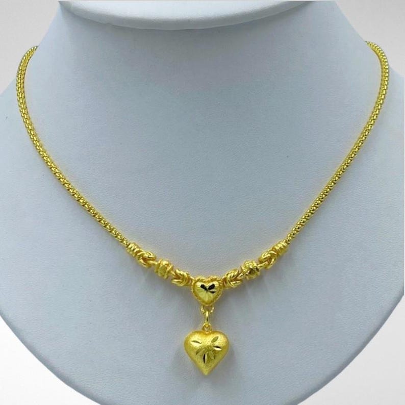 May include: A gold-colored necklace with a heart pendant. The necklace features a delicate chain and a heart-shaped pendant with a textured surface and a starburst design. The necklace is displayed on a white mannequin.