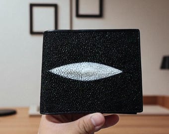 May include: A black bifold wallet crafted from textured stingray leather. The exterior features a prominent, elongated, diamond-shaped white patch of larger, smooth, pearl-like scales centered horizontally across the dark, granular surface.