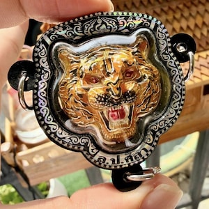 Puede incluir: Un colgante con forma de cabeza de tigre dorada con ojos rojos y boca abierta, engastado en un marco decorativo negro y plateado. El colgante tiene un reverso negro con anillos plateados.