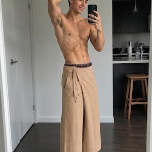 May include: Beige wide-leg trousers featuring a wrap-around waist design with a tie closure and a side pocket. The trousers have a textured, woven appearance and a straight-cut silhouette that falls to the mid-calf, paired with a dark patterned waistband.