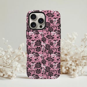 Puede incluir: Una funda de teléfono rosa con un diseño de encaje floral negro y lazos blancos. La funda tiene un acabado brillante. El diseño incluye rosas y lazos, creando una estética femenina.