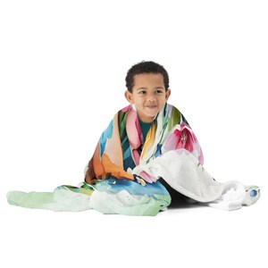 May include: A child wrapped in a blanket with a floral design. The blanket has a watercolour design with pink, orange, blue, and green colours. The blanket looks soft and cosy. The background is plain white.
