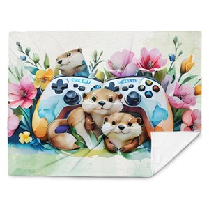 May include: A white blanket with a watercolour design. It features otters, game controllers, and flowers. The otters are brown and white, with smiling faces. The controllers are blue and white, and the flowers are pink, yellow, and blue.