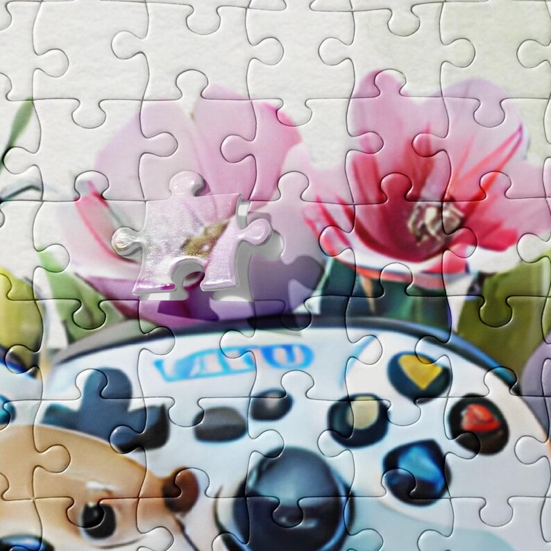May include: A jigsaw puzzle featuring a close-up of a white and black video game controller with blue and yellow accents. The puzzle also includes pink flowers and green leaves, creating a colorful and playful design.