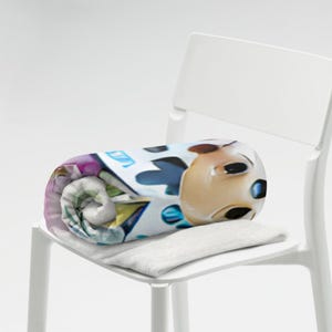 May include: Rolled blanket with a cartoon character design in tan, blue, and white. The blanket is on a white chair, with a small, rectangular, grey pillow underneath.
