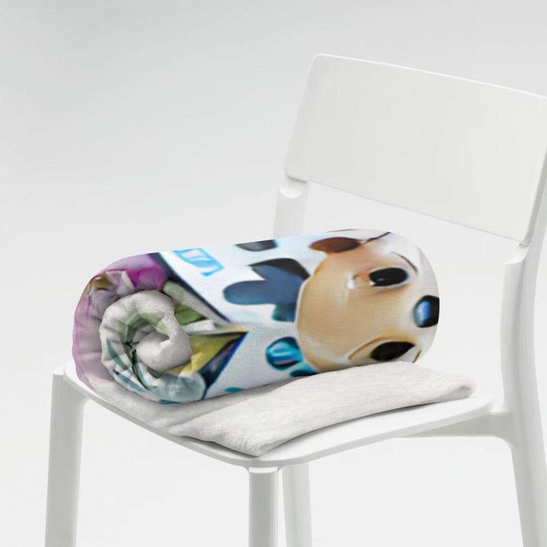 May include: Rolled blanket with a cartoon character design in tan, blue, and white. The blanket is on a white chair, with a small, rectangular, grey pillow underneath.