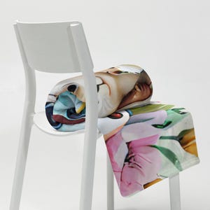 May include: A rolled-up throw blanket with a vibrant cartoon design, draped over a white chair. The blanket showcases a range of images, including a pink flower and a cartoon character. The blanket is made of soft material and is a decorative item.