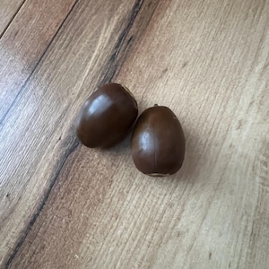 May include: Two smooth, dark brown acorns with a glossy finish rest on a light brown wooden surface. The acorns are oval-shaped with a small cap at the top. The wood grain provides a natural, rustic backdrop.