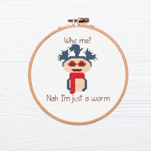 May include: A circular wooden embroidery hoop containing white fabric with a cross-stitch design of a blue-haired character wearing a red scarf. The text above reads Who me? and the text below reads Nah I'm just a worm in dark brown thread.