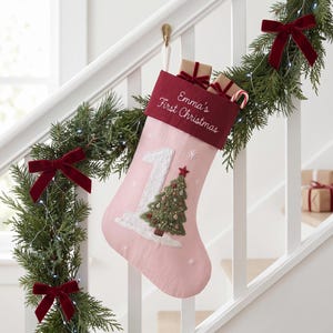 Può includere: Una calza natalizia rosa chiaro con un risvolto rosso scuro ricamato con il testo Emma's First Christmas. La calza presenta un grande numero uno bianco e un albero di Natale verde ricamato con piccoli ornamenti e una stella rossa.