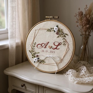 May include: Embroidered wedding hoop art featuring the initials "A & L" and the date "28.01.2023" in burgundy thread. The design includes floral accents and a geometric shape, set within a wooden hoop. A decorative piece for a wedding.