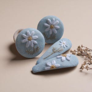 May include: A collection of light blue hair accessories, including two round hair clips and two barrettes. Each piece features a white daisy with a yellow center, embroidered on the fabric. The accessories are arranged on a neutral background.