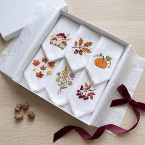 May include: A set of six white fabric napkins folded and arranged in a white gift box. Each napkin features distinct embroidery: mushrooms, oak leaves with acorns, a pumpkin, maple leaves, wildflowers, and red berries with autumn foliage.