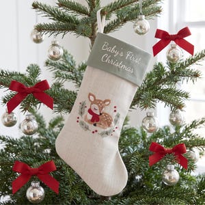 May include: A cream-coloured fabric Christmas stocking featuring a sage green cuff embroidered with the text Baby's First Christmas. The centre displays an embroidered fawn wearing a red scarf, surrounded by a wreath of green pine and red berries.