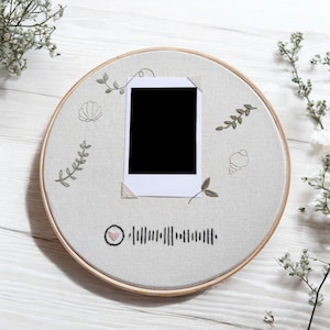 May include: A circular embroidery hoop in beige, featuring a blank polaroid photo. The fabric is adorned with embroidered leaves, shells, and a music barcode. The wooden hoop is set against a white wooden surface.