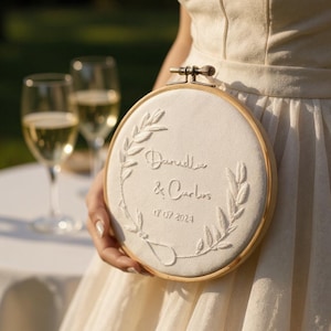 Puede incluir: Un aro bordado blanco con los nombres "Daniella & Carlos" y la fecha "17 07 2024". El bordado presenta un diseño de corona floral. El aro se sostiene frente a una persona que lleva un vestido blanco.