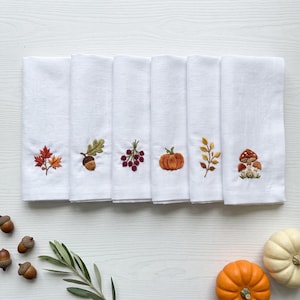 May include: A set of six white cloth napkins, each featuring a distinct embroidered autumn motif including maple leaves, an acorn, red berries, a pumpkin, a golden branch, and mushrooms, arranged in a horizontal row on a white surface.