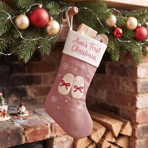 Peut inclure: Une chaussette de Noël rose poudré avec un revers blanc portant le texte brodé "Ava's First Christmas". La chaussette est décorée de flocons de neige blancs brodés et d'une paire de chaussons de bébé crème avec des nœuds rouges.
