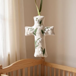 Puede incluir: Móvil de guardería en forma de cruz, de color crema, adornado con bordados de flores de lirio de los valles y hojas verdes. El móvil cuelga de una cinta verde. Fondo con una cuna de madera y una cortina transparente.
