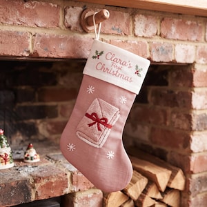 May include: A dusty rose Christmas stocking featuring a white cuff embroidered with the text Clara's First Christmas and holly sprigs. The stocking body displays a central embroidered baby blanket with a red bow and white snowflake accents.