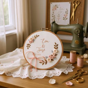 May include: A handcrafted embroidery hoop featuring the names "Eva & Jose" and the date "2 07 2023" in elegant script. The design includes floral and leaf accents in soft pinks and browns, with a delicate pink ribbon. The hoop is displayed on a white lace-trimmed cloth.