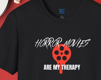 May include: Black t-shirt featuring the text "HORROR MOVIES ARE MY THERAPY" in white, with a red film reel graphic. The shirt has a crew neck and short sleeves. The brand logo "Feral Blue Designs" is visible.