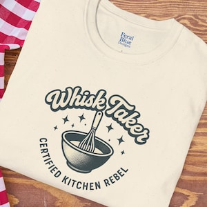 May include: Cream-colored crew neck t-shirt with the text "Whisk Taker" above a bowl and whisk graphic. The words "Certified Kitchen Rebel" are printed around the bowl. The shirt has a speckled texture and the brand name "Feral Blue Designs" is printed on the upper right.