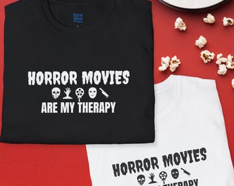 May include: Black and white t-shirts with the slogan "HORROR MOVIES ARE MY THERAPY" and horror-themed graphics. The shirts are on a red surface with popcorn and a film reel.
