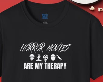 May include: Black t-shirt featuring the white text "HORROR MOVIES ARE MY THERAPY". The design includes skull, film reel, and mask graphics. The shirt has a crew neck and short sleeves.