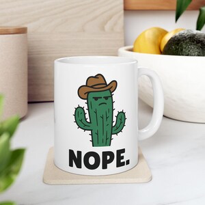 Può includere: Tazza in ceramica bianca con un cactus dei cartoni animati con un cappello da cowboy marrone e la parola "NOPE." in nero. Il cactus ha un'espressione scontrosa. La tazza è su un sottobicchiere beige.