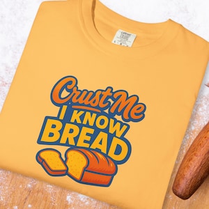 May include: Mustard yellow t-shirt with the text "Crust Me I Know Bread" in orange and blue. The design includes illustrations of bread slices and a loaf of bread. The shirt has a Comfort Colors tag.