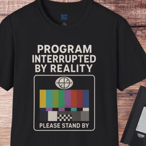 May include: Black t-shirt with the text "PROGRAM INTERRUPTED BY REALITY" above a colorful test pattern graphic. The graphic includes the words "PLEASE STAND BY". The shirt also has a small logo in the upper left corner.