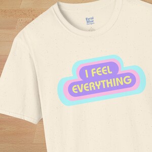 May include: Cream-colored t-shirt with the text "I FEEL EVERYTHING" in a colorful cloud design. The text is in yellow, purple, and blue. The shirt has a crew neck and short sleeves. The brand name "Feral Blue Designs" is printed on the shirt.