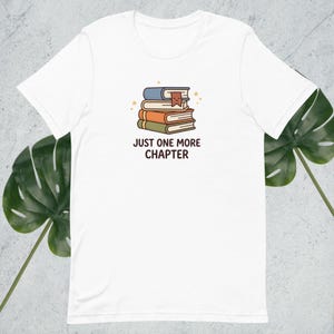 May include: White t-shirt featuring a graphic of three stacked books with a bookmark and the text "JUST ONE MORE CHAPTER" in black. The top book is blue, the middle is orange, and the bottom is green.