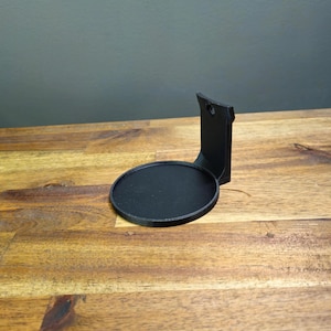 May include: A black, circular wall mount for a speaker or other device. The mount has a flat, round base and a vertical support with a hole for mounting. The mount is made of a matte black material and sits on a wooden surface.