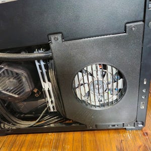 May include: Close-up of a black computer case with a cooling fan visible. The fan is behind a metal grill. Other components, including a water cooler and wiring, are also visible inside the case. The case has a textured surface.