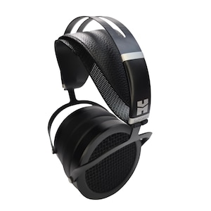 Puede incluir: Auriculares circumaurales negros con diadema de cuero texturizado, marco de metal y copas circulares con rejilla de malla fina. El lateral de la diadema muestra el logotipo de Hifiman y el texto SUNDARA en letras plateadas sobre una base negra.