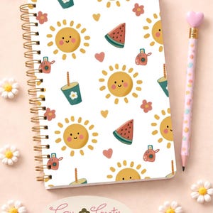 May include: A notebook with a summer pattern of smiling suns, watermelon slices, cameras, and drinks. It has a gold spiral binding and a pencil with a heart-shaped eraser. The text "Cau Creates" is visible. The notebook is surrounded by small white flowers.