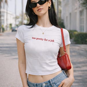 May include: White crop top with the text "too pretty for a job" in red. The shirt is paired with blue jeans and a red shoulder bag. The person is wearing black sunglasses and gold hoop earrings.
