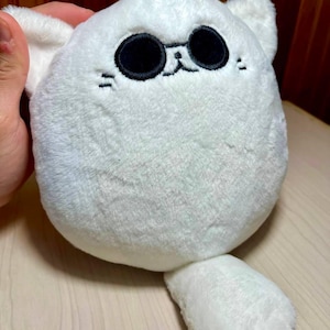 May include: A white plush toy shaped like a cat with large black eyes and small embroidered whiskers. The cat has two small ears and a single paw extending from its body. The toy is soft and fluffy.