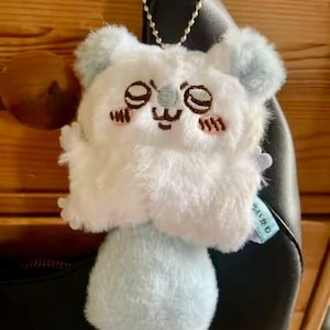 May include: A plush keychain in the shape of a white and light blue cartoon character with closed eyes and embroidered details. The keychain is attached to a silver chain.