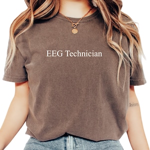 May include: A brown t-shirt with the text "EEG Technician" in white. The shirt is a neutral color and has short sleeves. The model is wearing a gold necklace and blue jeans.
