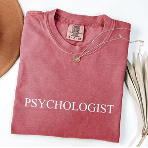 May include: A dusty rose-colored t-shirt with the word "PSYCHOLOGIST" printed in white. A gold-colored necklace with a pendant rests on the shirt. The shirt is folded, and a brown hat and dried leaves are visible.