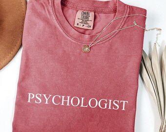 May include: A dusty rose-colored t-shirt with the word "PSYCHOLOGIST" printed in white. A gold-colored necklace with a pendant rests on the shirt. The shirt is folded, and a brown hat and dried leaves are visible.