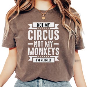 May include: A brown t-shirt with the text "NOT MY CIRCUS NOT MY MONKEYS I'M RETIRED!" in white. The shirt has short sleeves and a crew neck. The text is arranged in a bold, eye-catching design.