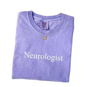 May include: A folded, light purple t-shirt with the word "Neurologist" printed in white. A gold-colored necklace with a pendant rests on the shirt. The shirt has a crew neck and the brand "Comfort Colors" is visible.