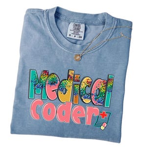 May include: A light blue Comfort Colors t-shirt with the words "Medical Coder" in colorful, outlined letters. A gold necklace with a pendant rests on the shirt. The shirt has a small tag at the neckline.