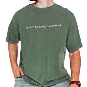 May include: A faded green t-shirt with the text "Speech-Language Pathologist" in white. The shirt has a crew neck and short sleeves. The person wearing the shirt is also wearing blue jeans and a brown watch.